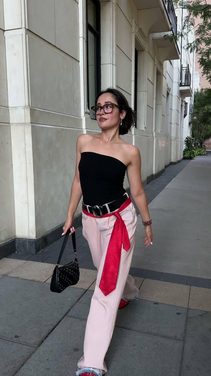 Bustier noir et pantalon rose, foulard rouge noué à la taille, sur un trottoir bordé d'immeubles clairs.